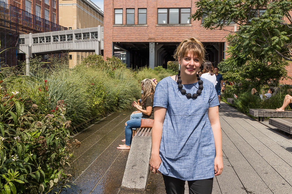 A woman is smiling and facing the camera. She has white skin and long, straight blond hair in a ponytail and bangs that cover her forehead. She is wearing a blue denim shirt with short sleeves, black pants, and a black necklace made of large circles that goes down to her chest. Around her are garden foliage and a walking path made of gray cement. In the background is the ground entrance to a building and a pathway to the left of it. 