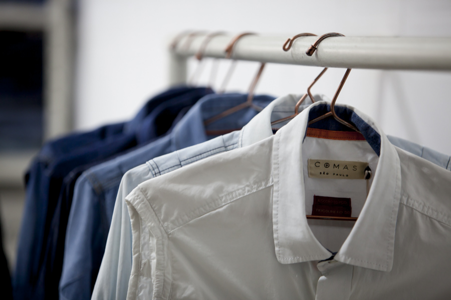Shirts hanging on a metallic rack, on a gradient from white to dark blue, with the white shirt closest to the camera, displaying a beige label with the brand "Comas - São Paulo" written in black. In the background is a white wall.