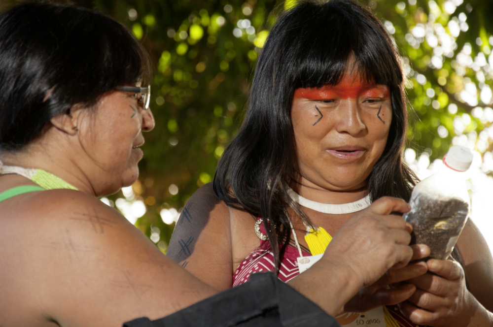 Two indigenous women are having a conversation. The woman on the left of the photo is in profile, pointing to an object in the other woman’s hands, who is looking at it and facing towards the camera. The woman on the left is wearing black-rimmed glasses and has straight black hair that is cut-off by the photo’s left margin. The woman on the right also has long, straight black hair with bangs that cover her forehead. She is wearing a white necklace, and has a red band of paint across her face that goes over her eyelids and eyebrows. Below each of her eyes is a drawing of half a diamond shape with a dot at the center and the tips facing inwards toward her nose. She is holding a small, clear plastic bottle filled with earth and with a white cap.