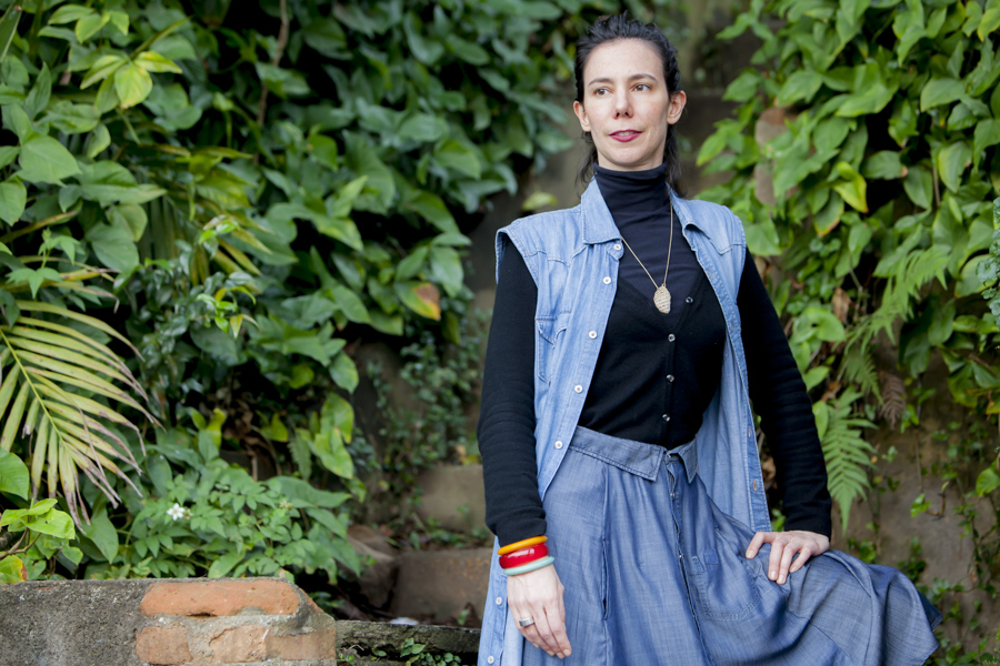 A woman is facing the camera, looking towards the left side of the photo, resting her left hand on her left thigh. She has white skin and straight dark brown hair, tied back. She is wearing a light jeans skirt, a black turtleneck shirt under light jeans overalls, colorful bracelets on her right wrist, and a gold string necklace with a beige pendant that hangs near her chest. In the back is a wall covered in foliage. 