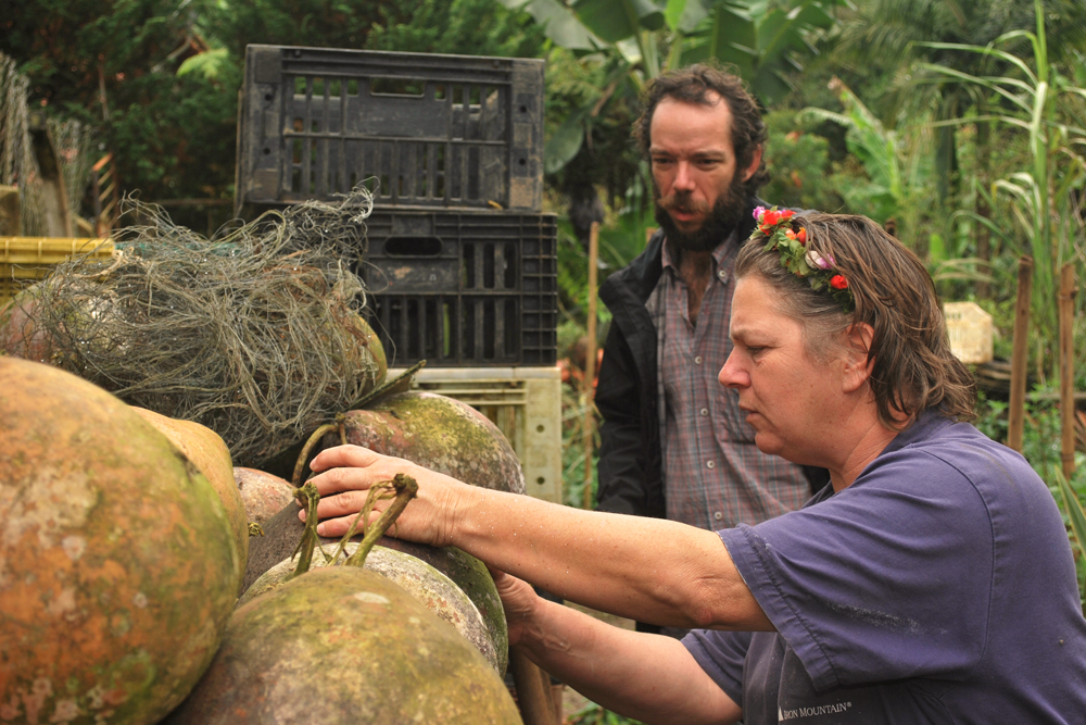 The same man from the cover photo, with white skin, beard and moustache, and short, wavy light brown hair appears in the background towards the right, facing the camera diagonally, looking at a row of large green gourds. Next to him, closer to the camera, is a woman in profile handling the gourds. She has white skin, short, straight light brown hair with a garland of small red flowers. She is wearing a short-sleeved blue shirt and has a serious expression on her face. In the back are stacks of black plastic crates. Further back is some tall, dark green foliage.