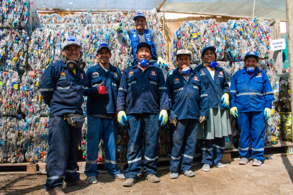 Men and women in uniform, wearing dark blue caps, pants and long-sleeve shirts with thick work gloves pose for a photo. They are also wearing blue face masks, hanging by their necks, and some of them are smiling. All have bronze skin and dark hair. The ground is made of gray cement. Behind them are piles of recyclable plastic material stacked on wooden pallets.