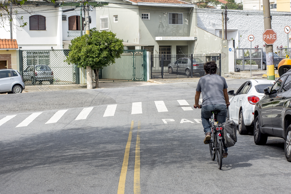 The same man from the cover photo, with white skin, thin beard and short, wavy dark brown hair, is riding a bicycle on a street towards the back of the photo. Ahead of him is some white signage on the asphalt, written "Stop" in Portuguese, and a pedestrian crossing. To the left are some parked cars. Further back are some gated houses and a short tree.