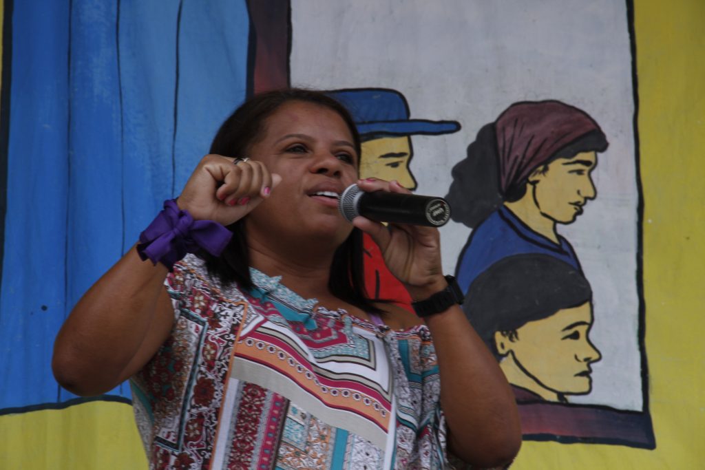A woman, facing diagonally towards the right corner of the photo, talks into a microphone. She holds the microphone with her left hand and has her right arm up with her fist closed. The woman has dark skin, straight dark brown shoulder-length hair, and wears a short sleeve shirt with colorful print. On her left wrist is a watch with a black band. On her right wrist is a purple band tied into a knot. Behind her is a yellow banner with a drawing of an open window with the faces of workers in profile. 