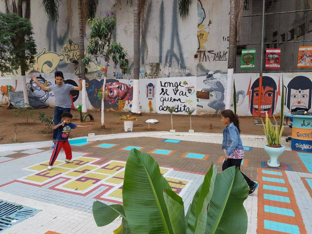 Two children and a young adult play on a hopscotch board painted onto the ground in a park. The young adult and the boy are on the left side of the photo, and the girl is on the right. The boy is stepping on the numbers 1 and 2 of the hopscotch board, while the girl is watching him. All three are wearing colorful clothes. In the back is a wall with graffiti of faces and the phrase “Those who believe will go far” written in Portuguese.
