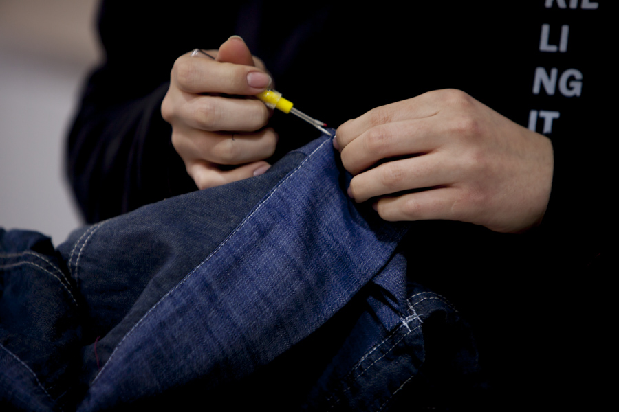 A white person's hands are handling some dark jeans with a sharp metal tool that is about the size of their fingers. In the background is the person's torso, wearing a black sweatshirt with some white letters sewn into the chest.