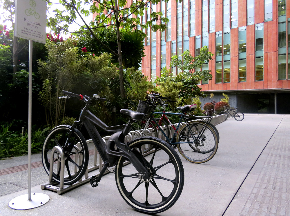 The same black bicycle from the cover photo appears parked in a bike rack next to other bikes, facing diagonally to the left and the back of the photo. The bike rack is silver and is on top of a gray cement floor. To the front of the bicycles, in the left corner of the photo, is a garden with various small trees. In the back is a building with walls covered with brick-colored tiles, with tall, vertical rectangular glass windows.