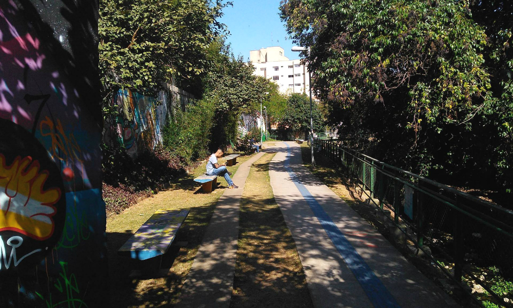 A person with a shaved head is sitting on a bench, their legs crossed, and looking down at the ground. They are wearing jeans and a gray t-shirt, the sun is shining on them between the shade of the surrounding trees. Cutting through the center of the photo is a gray concrete walking path, passing in front of the bench and towards the back of the image, with a blue tactile floor and short grass on either side. There are trees at the edges of the photo, and in the back are buildings and a clear, blue sky.