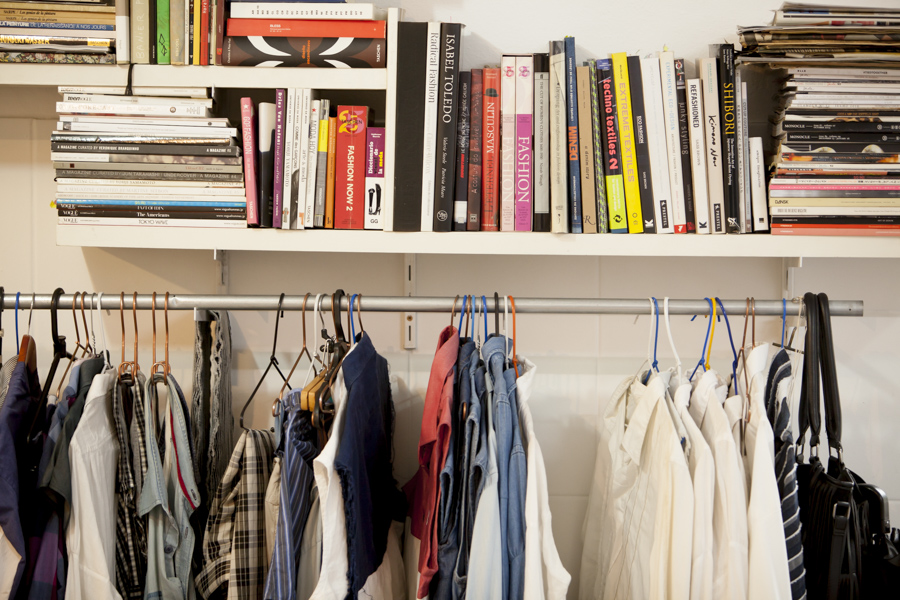 A foto mostra uma parede branca com uma estante, virada para a câmera, cheia de livros. Abaixo da estante, há peças de roupas penduradas em cabide, ao longo de um tubo metálico prateado.