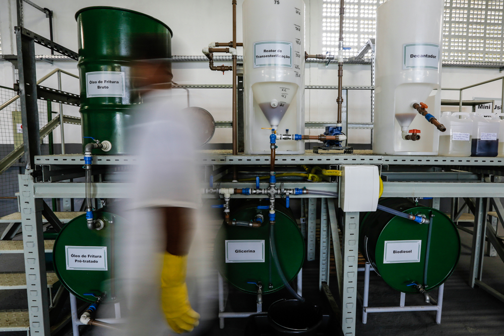 A person walks past dark green barrels and large, clear white containers in an industrial laboratory. The person is wearing white clothes and yellow rubber gloves, and their image is completely out of focus. 