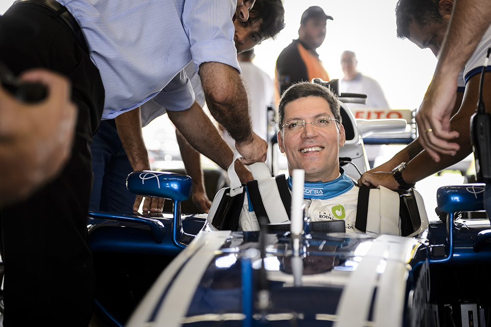 A man smiles while sitting in the driver’s seat of a racecar. The hands of other men are on his shoulders, helping attach white safety belts. The racecar is white, as is the man’s uniform, and he is wearing rectangular wireframe glasses. He has white skin and short, straight brown hair. In the back is the out of focus image of other men and a gray sky.