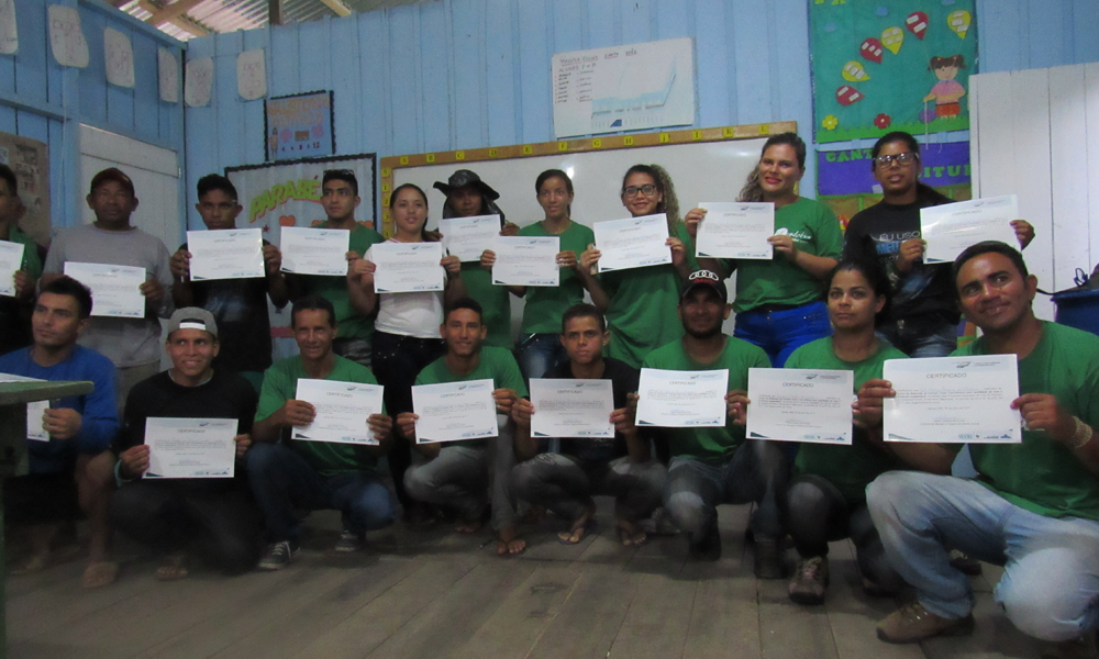 People wearing identical green t-shirts as a uniform pose for a photo holding up certificates. Behind them is a light blue wooden wall, with a whiteboard hanging on it along with some posters that are out of focus. The floor is made of dark wood.