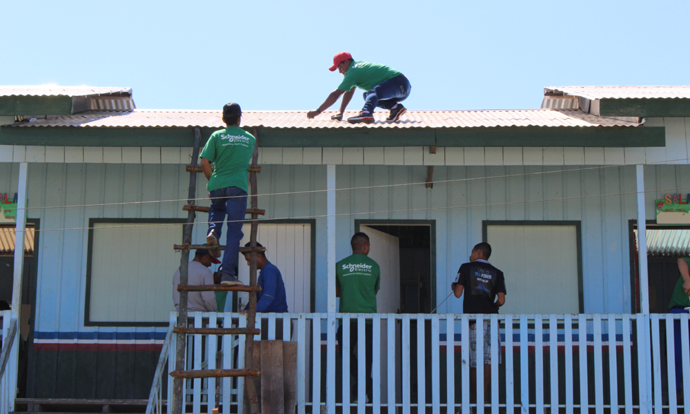 Men work on installing solar panels on a one-floor house with white wooden walls. One of the men is on the roof, squatting down and facing the left side of the photo. There is another man climbing up a rustic wooden ladder, towards the rooftop, with his back to the camera. The others are spread out around the area. Most of them are wearing identical green t-shirt as a uniform. The house has a white wooden fence on the edge of its front porch. In the background is a clear blue sky.