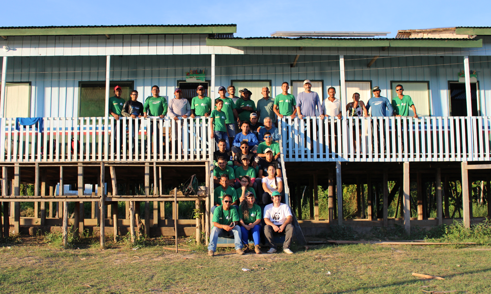 Trabalhadores uniformizados com camisetas verdes posam para a foto, preenchendo a escada e parte da sacada de uma casa suspensa de madeira branca. O chão é de grama baixa. Ao fundo, o céu azul ensolarado, sem nenhuma nuvem.