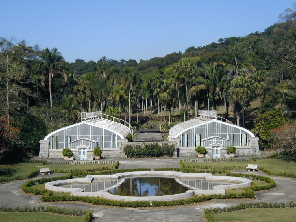 The photo shows two white architectural structures made of glass and metal, shaped into triangular domes. In front of them, closer to the camera, is a reflecting pool with white borders, surrounded by a gray cement walking path, which is further surrounded by small, trimmed bushes. In the background are palm trees and other tall trees. 