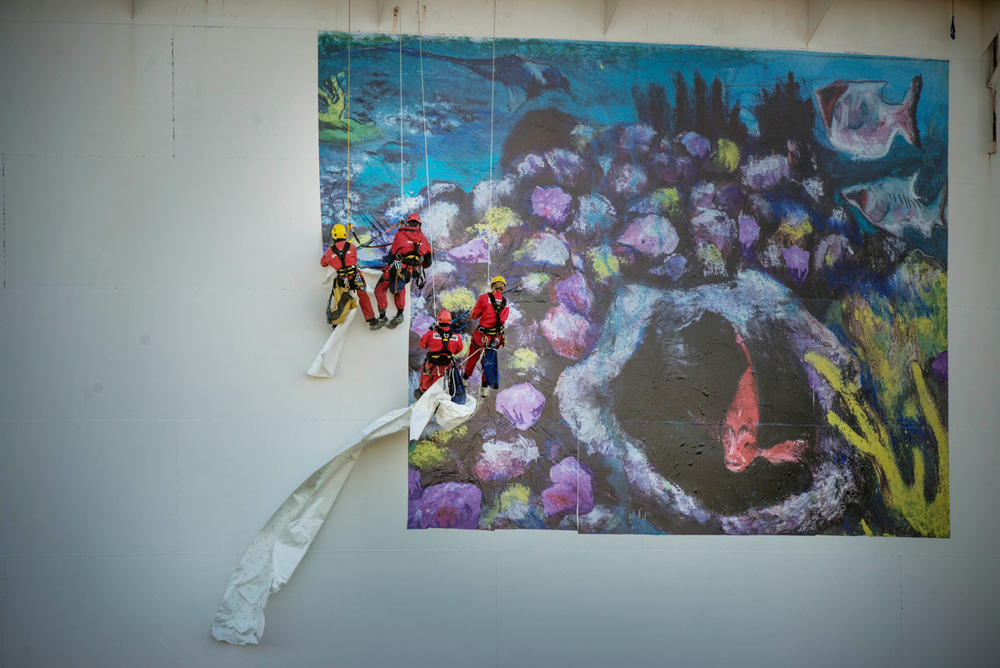 Quatro trabalhadores vestidos com macacões vermelhos e capacetes vermelhos ou amarelos, estão pendurados por cordas aplicando uma grande ilustração com tecidos sobre um paredão branco. A ilustração mostra o oceano repleto de peixes coloridos e corais.