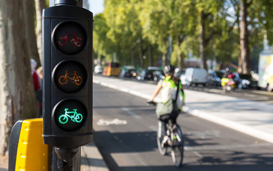En primer plano, y en el lado izquierdo de la foto, un semáforo con iconos en forma de bicicleta tiene la luz verde iluminada. Al fondo, un ciclista pedalea en una ciclovía hacia el fondo de la foto. En el lado derecho de la imagen se ve la calle con carros, con un cierto tráfico, y más a la derecha, una hilera de árboles altos.