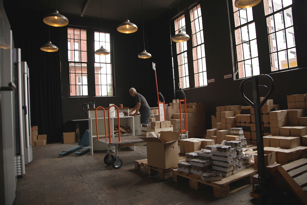 A man with his back to the camera is working inside a warehouse. Around him are closed cardboard boxes of various sizes, and shaded walls with large rectangular glass windows with dark wood frames. The floor is dark gray cement. At the top of the photo are light fixtures in a flattened bell shape hanging by wire with their lights on.