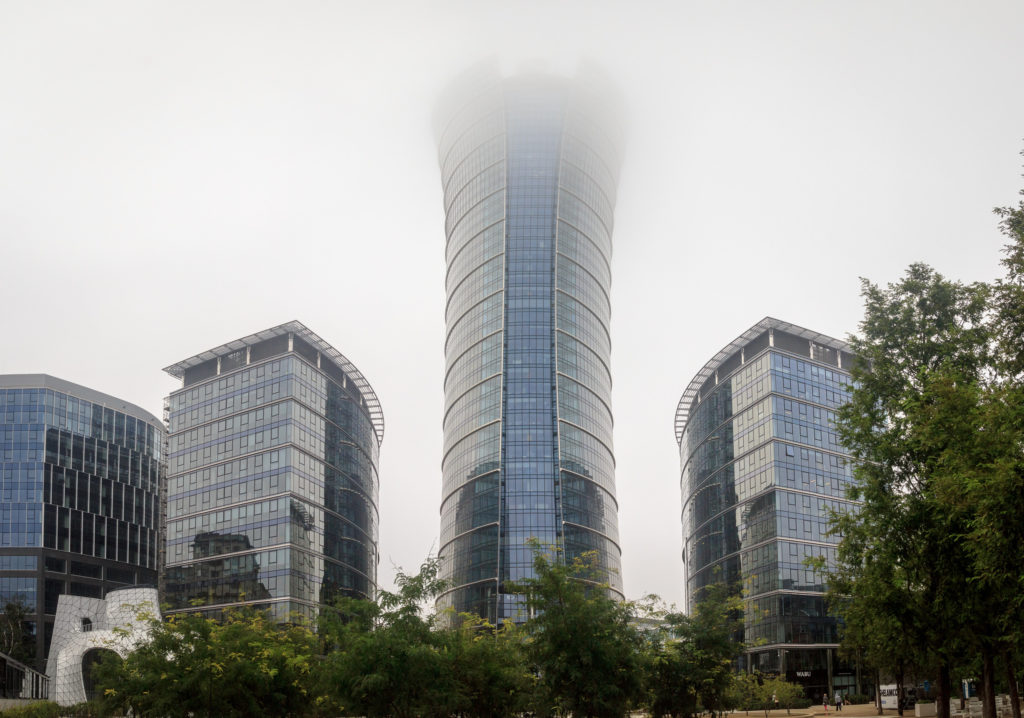 The photo shows four glass buildings with silver and grayish blue tones. The leftmost building, cut-off at the edge of the photo, is the lowest, with a plain surface and irregular shape. To its right is a slightly taller semi-cylindrical building, with a flat side facing diagonally towards the left of the photo. At the center is a tall building that reaches the photo's upper edge, with a cylindrical shape that opens up slightly at the top. This building has a vertical strip of blue glass, while silver glass covers the rest of its exterior. Finally, to the right is a building identical to the previously mentioned semi-cylindrical one, but with its flat side facing towards the right. In front of the buildings are some small trees with dark green leaves. The sky in the back is cloudy.
