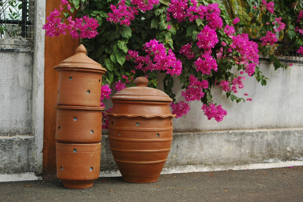 A bouganvillea bush with bright pink flowers grows over a white wall with white grating. The bush's branches hang down close to the ground, where there are three orange clay pots in a stack and, to the right of them, a larger pot made from the same material. The objects are round with holes. They are resting on the ground.