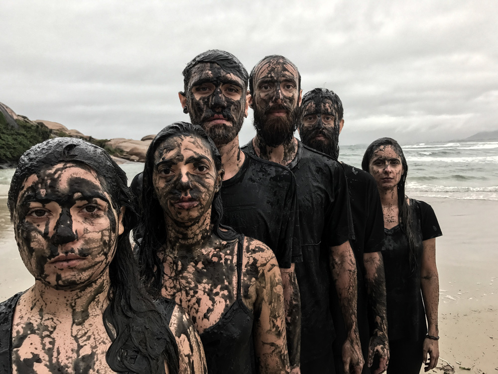 Six young people, wearing black t-shirts and tank tops, look at the camera, their faces and bodies covered in black oil. All of them have white skin and straight brown hair. In the background is the sea and a cloudy sky.