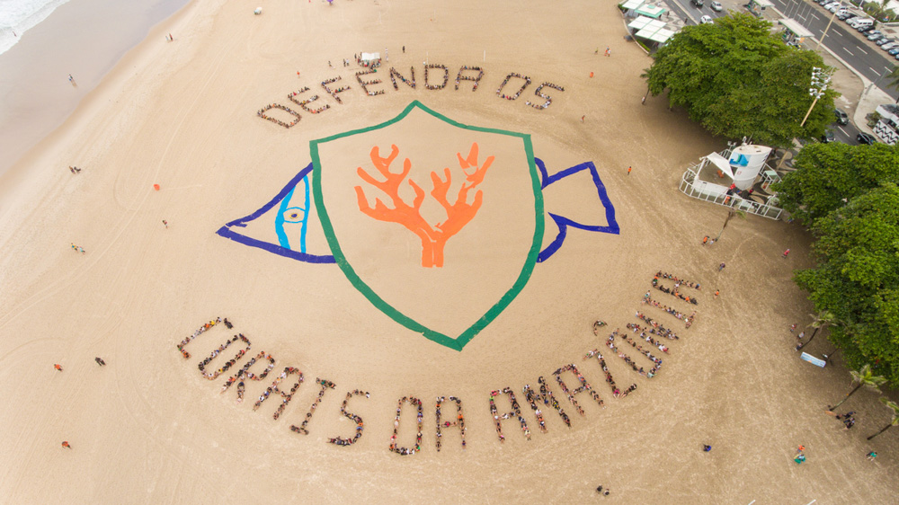 Os dizeres “Defenda os corais da Amazônia” são formados por inúmeras pessoas organizadas na areia de uma praia, vista de cima, em torno da ilustração de um peixe azul, com um escudo verde no qual está a silhueta de um coral cor-de-laranja. 