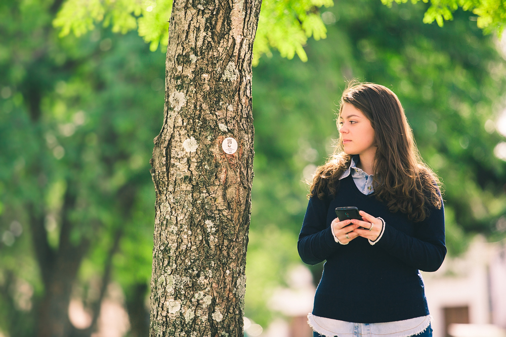 The same tree trunk from the previous photograph, with the small circular plaque containing a QR code, is seen from a distance in the center of the photo. To the right, a young white woman with long, straight brown hair holds a cell phone and looks at the plaque. The woman is wearing a light blue shirt under a dark blue sweater. In the background is out of focus foliage. 