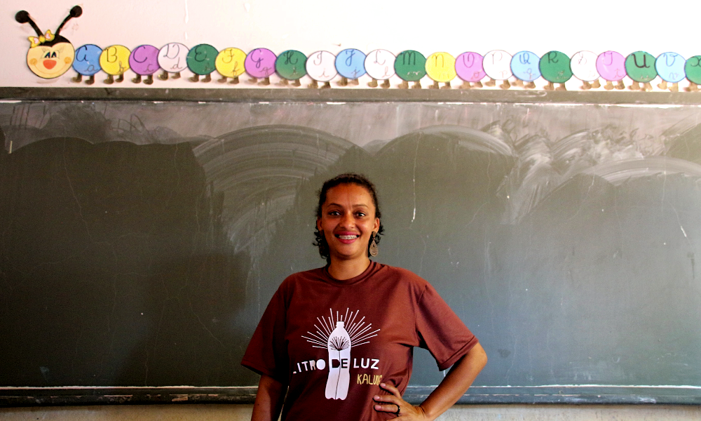 A woman smiles at the camera, her left hand resting on her hip. She has black skin and dark curly hair tied into a ponytail. She is wearing brown earrings shaped like water drops and a dark brown t-shirt with the “Litro de Luz” logo printed in white on the chest. Behind her is a green chalkboard, above which is glued a colorful paper caterpillar with the letters of the alphabet printed on each section of its body. The wall is white.