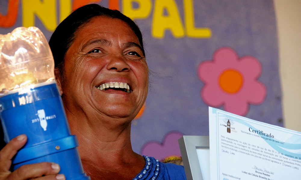 A woman smiles, facing diagonally towards the right side of the photo. She is holding a plastic bottle lamp in her right hand, and a certificate printed on white paper in her left hand, with the word “certificate” written on it in Portuguese in dark blue and the “Litro de Luz” logo printed in brown. The woman has reddish skin and straight black hair, tied back. In the back is part of a large purple poster with yellow letters.