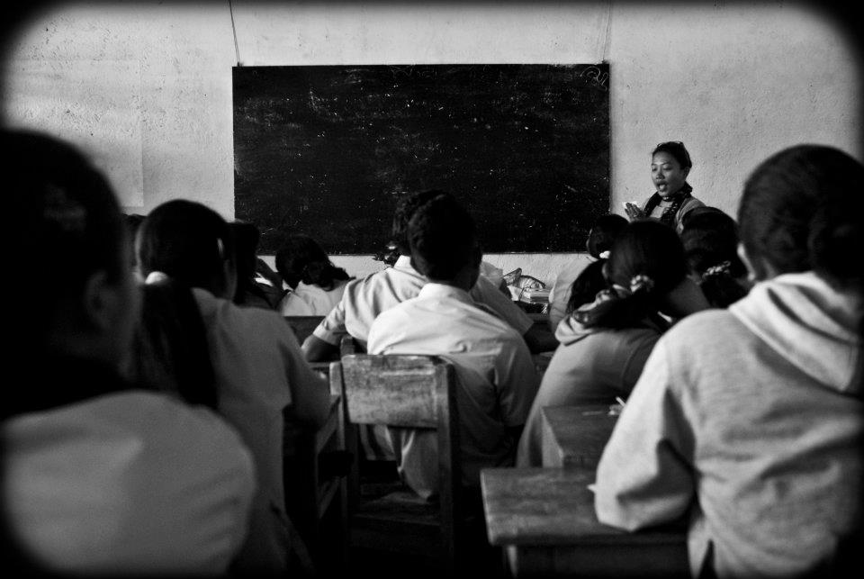 A black and white photo shows children sitting in rows on wooden chairs, their backs to the camera. In front of the children appears a standing woman's face, with a talking expression. The woman has dark skin and dark hair, tied back. In the background is a white wall with a large blackboard hanging on it.