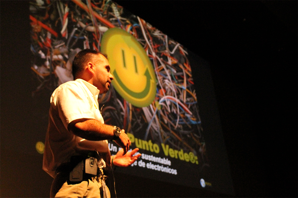 Foto de Alvaro en un escenario, haciendo una conferencia. Al fondo, una pantalla mostrando un emoji feliz y escrito "Punto Verde". En la foto, vemos a Alvaro, vistiendo una camisa blanca, de perfil, de la cintura hacia arriba