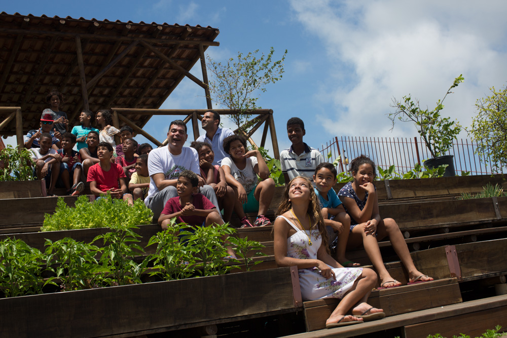 Na foto, vemos Demetrius com um grupo de jovens e crianças, sentados na escada do telhado ecoprodutivo. Ao lado, um canteiro com várias verduras. Todos estão sorrindo e olhando para a câmera. 
