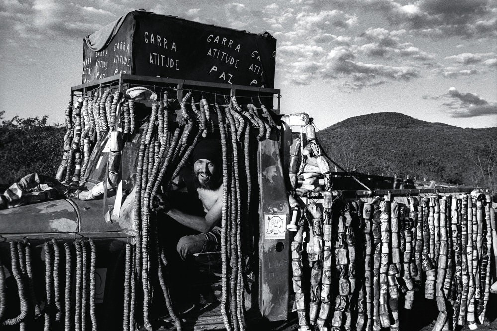A black and white photo same man from the cover photo sitting in a truck, opening with his left hand across the front of his body a curtain made of round objects that covers the left door of the automobile, and smiling at the camera with his mouth open. He is wearing a black cloth over his hair, is shirtless, and wears dark pants. The truck's exterior is covered entirely by curtains made of string with vertical rows of aluminum cans and other material hanging on them. On top of the truck's cabin is a rectangular structure covered by a dark cloth, on which are written many times the words "tenacity, attitude, peace" in Portuguese in white letters. In the back is a valley and the sky with some clouds. 