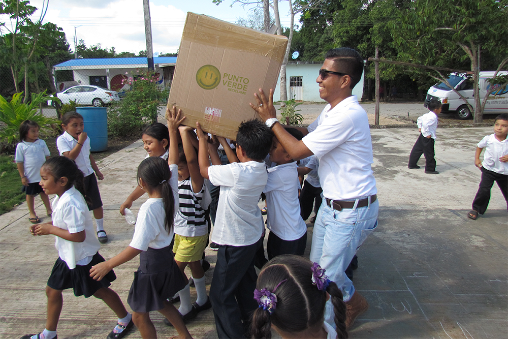 A man, medium height, carrying a large cardboard box. With him, a group of several children under 10 years old, helping to carry the box
