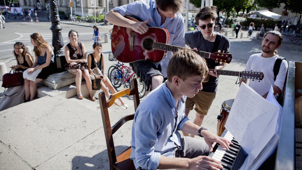 A young man is playing a piano on the sidewalk. Two other young men are playing guitar and another young man watches them closely. A few steps back, four women sitting on the sidewalk are watching the musicians.
