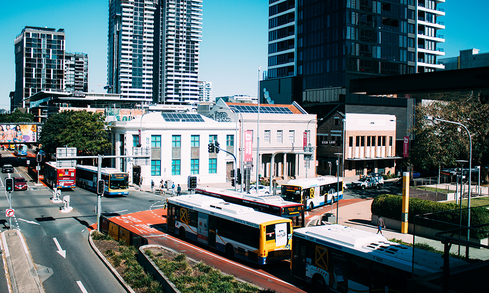 A foto mostra o centro de Brisbane, em um cruzamento com vários ônibus e prédios. Dois deles, de dois andares, estão com placas fotovoltaicas. 