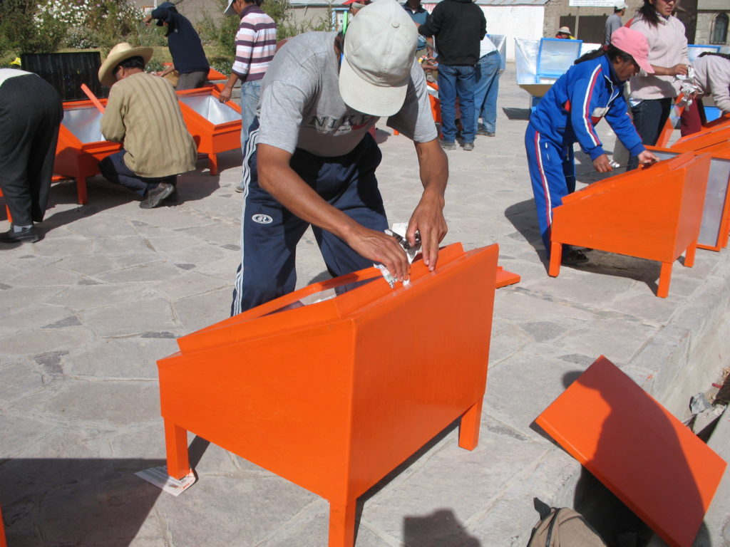 The picture shows a group of more than ten people. They are putting the final touches on solar cookers they have made. In the foreground is a man of medium height, wearing a cap, with his head down (we cannot see his face). He is varnishing the solar cooker (a wooden box with a glass lid, approximately one square meter in size).