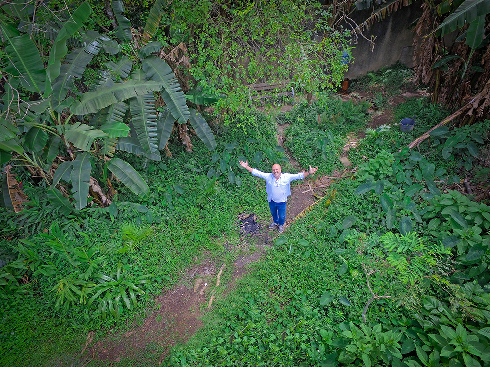A foto mostra uma imagem aérea, em que vemos um homem olhando para cima com os braços abertos, em um espaço cheio de verde, árvores e plantas.