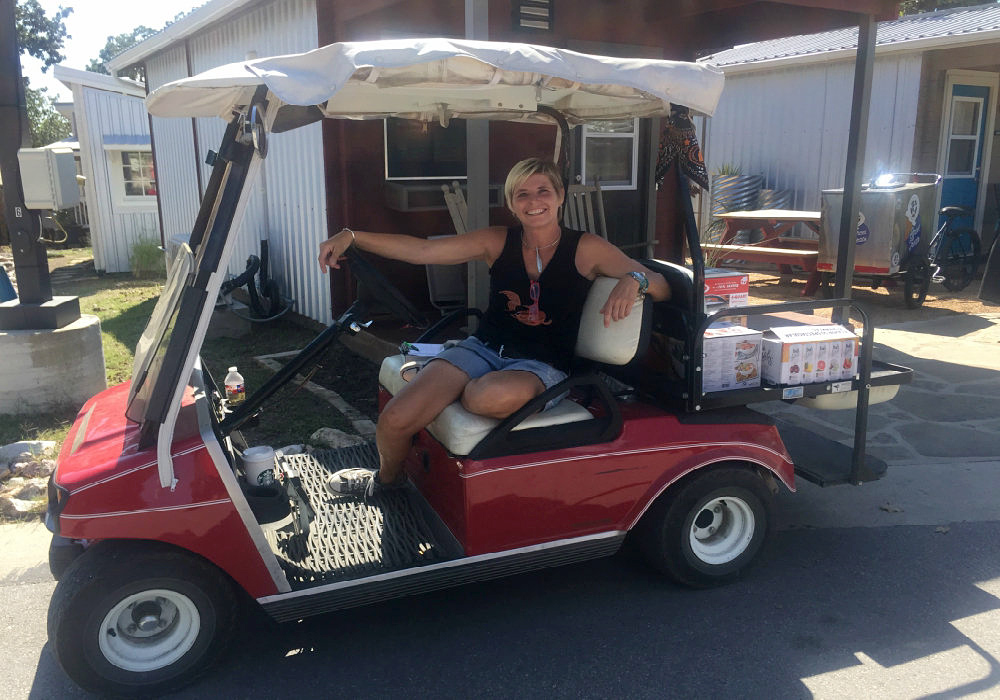 A young, blonde, short-haired woman is smiling at the camera. She is in a red golf cart, which is open and could seat two passengers. A small wooden house is in the background.
