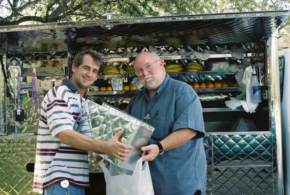 A white man around the age of 40 is wearing a colorful striped shirt. He is smiling at the camera and holding some kind of laminated box. On the right side is a balding man, older than the first one. They are helping each other with the box.