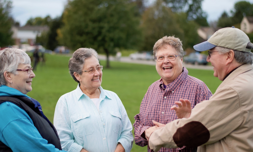 Four people with short graying hair, all wearing glasses, are talking and smiling. In the background, a grassy field, some trees, some houses and a street. Some cars, too blurry to make out clearly, are parked on the street. 