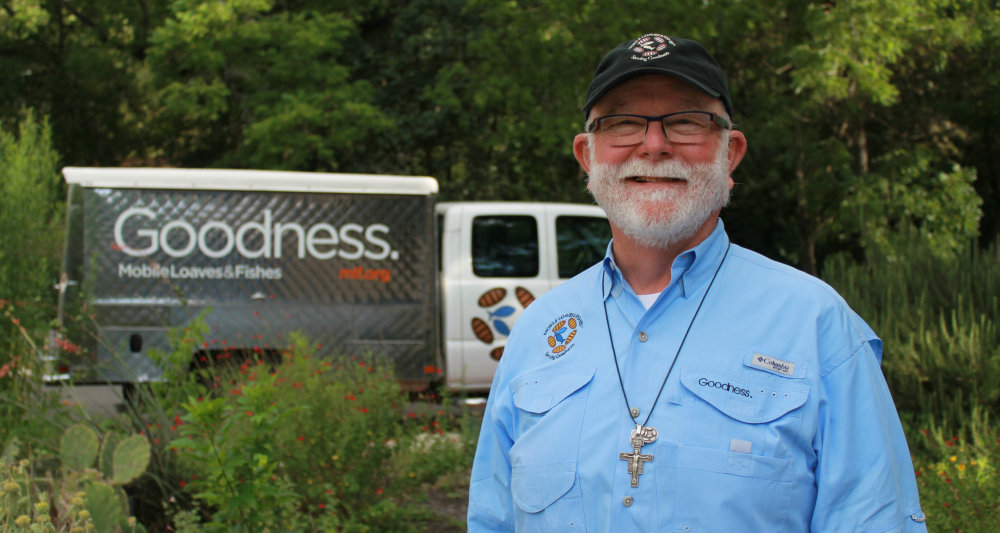 Um homem idoso, branco, com barba branca, vestindo uma camisa azull, óculos e boné preto. Ele sorri para a câmera. Atrás, um caminhão com a palavra "Goodness" (bondade).