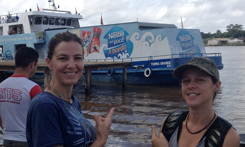 A thin white woman with her brown hair up is in the left side of the picture, smiling at the camera. Standing next to her, another thin white woman, shorter than her companion, is wearing a black cap and a backpack. She also smiles at the camera. Both are pointing at a boat in back of them, docked in muddy waters, decorated with the Nestlé logo and pictures of Nestlé products.