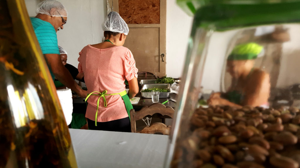 As if the camera is on the back wall of a shelf, a glass bottle is to the left, and part of a glass jar of beans is on the right. Both objects are blurry. In the kitchen, a woman and man, both wearing white caps on their heads, have their backs to the camera. They are cutting vegetables. Next to them, through the jar of beans, another woman, her image blurred by the glass, wears a green cap. She seems to be peeling some vegetables.