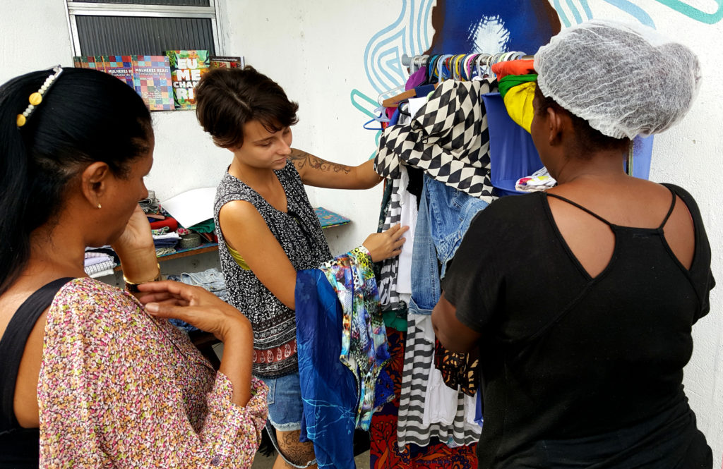 Uma mulher branca, cabelo negro e curtinho, magra, está mostrando uma arara com várias roupas coloridas para outras duas mulheres, negras, que estão de costas para a imagem. 