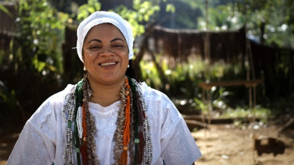 A black woman wearing a white turban, a white blouse and several colored beaded necklaces smiles at the camera. She is standing in a yard with lots of plants.