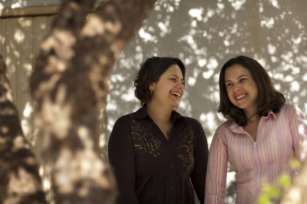 Two women are standing side by side, in the right side of the picture, laughing together without looking at the camera. The first woman (left) has white skin, and is shorter than her companion, with wavy hair. She is wearing a black shirt. The woman to her right has shoulder-length dark hair and is wearing a shirt with pink vertical stripe. On a white wall behind them are the shadows of tree leaves. Part of a tree trunk is out of focus. 