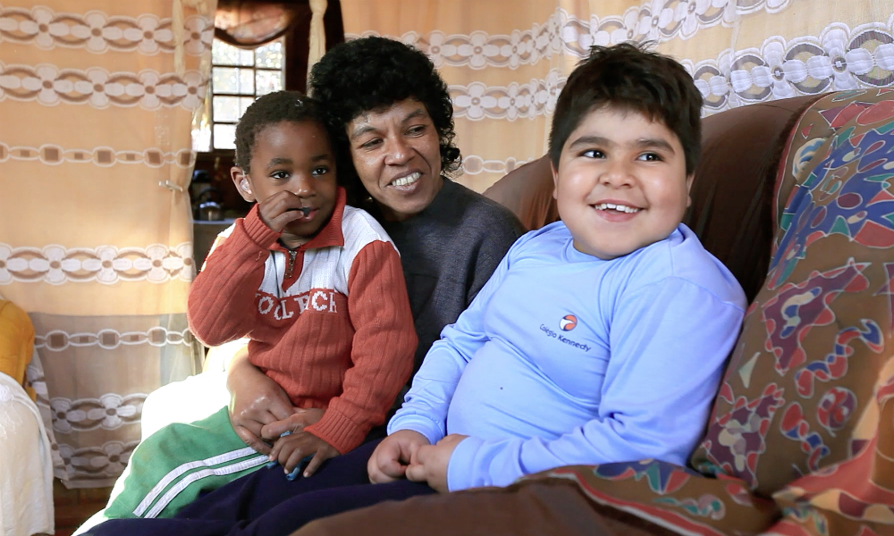 A black, curly-haired woman is sitting with a little boy, also black and slim, on her lap on a brown couch. Next to them on the couch is another boy, who is very chubby, with lighter skin, brown hair and brown eyes. Both the mother and the bigger boy are smiling,. She is looking at him, and he is looking at someone who seems to be behind the camera.
