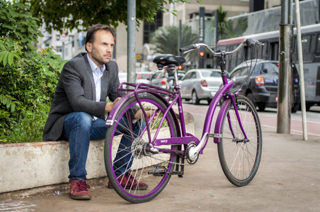 The same man featured in the cover photo, wearing jeans, a light blue shirt, a dark gray blazer and burgundy shoes, is sitting on the edge of a bed of lush greenery on a busy avenue. There is a purple bike in front of him. 
