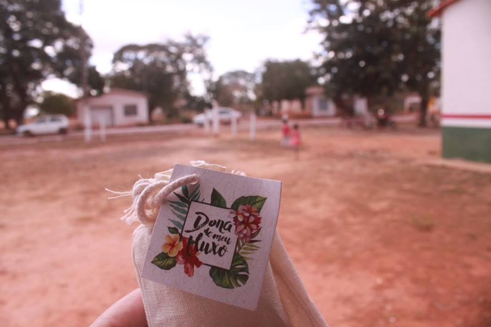 In the foreground, part of a white hand holds a small beige fabric bag, tied with a string. On the bag there is a small, square, paper card decorated with an image of flowers and the inscription: "Owner of my Menstrual Flow." In the background is sandy soil, some small houses and a couple cars, out of focus. 